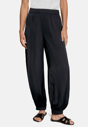 Femme portant un pantalon noir ample à taille élastique avec des poignets élastiques, associé à des sandales noires à bouts ouverts et un haut blanc, debout devant un fond uni.