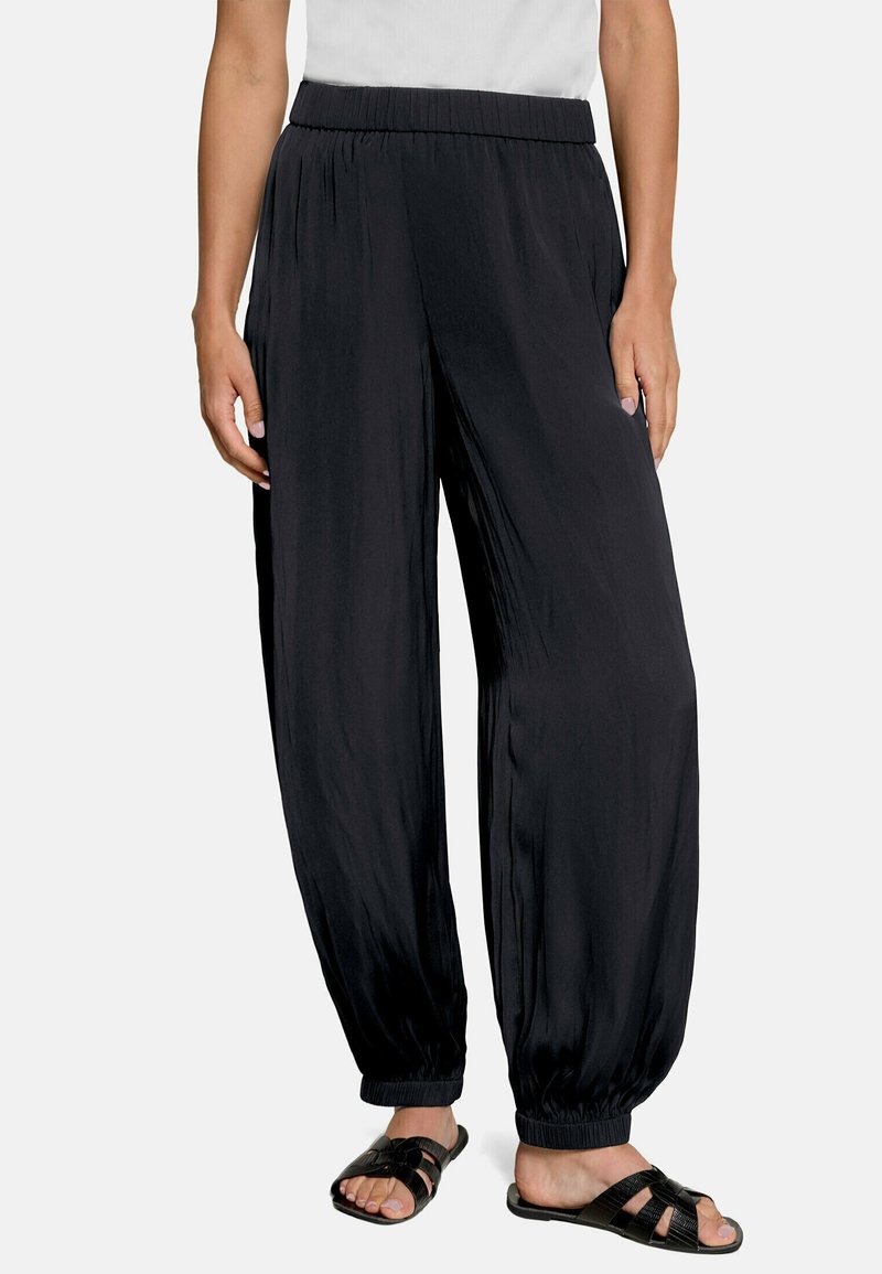 Femme portant un pantalon noir ample à taille élastique avec des poignets élastiques, associé à des sandales noires à bouts ouverts et un haut blanc, debout devant un fond uni.