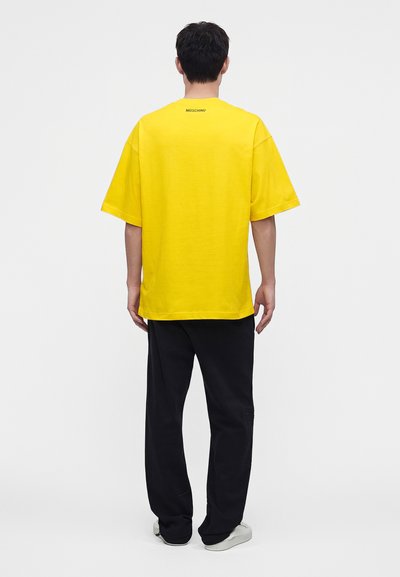 Άτομο που φοράει ένα φωτεινό κίτρινο oversized t-shirt με μαύρο παντελόνι και λευκά αθλητικά παπούτσια, στέκεται με την πλάτη στραμμένη σε ένα απλό φωτεινό φόντο.