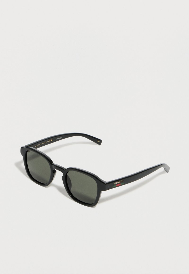 Lunettes de soleil Gucci à monture carrée noire avec verres foncés et logo sur les branches, posées sur un fond blanc.