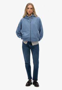 Lichtblauwe hoodie met rits aan de voorkant, twee voorzakken en een koord. Draagt over een verticaal gestreept shirt en gecombineerd met donkere jeans.