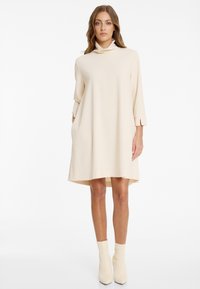 Frau, die ein cremefarbenes langärmliges Rollkragenkleid mit Taschen und passenden Stiefeletten trägt, steht vor einem schlichten weißen Hintergrund.