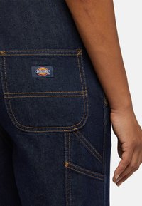 Dark blue denim overalls with contrast orange stitching, featuring a back pocket and a branded label. The fabric has a smooth, sturdy texture.
