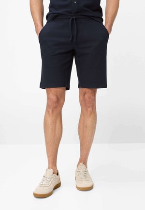 Uomo con pantaloncini blu navy con coulisse e tasche laterali e sneakers beige, in piedi davanti a uno sfondo bianco con le mani nelle tasche.