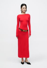 Czerwona dopasowana sukienka z długim rękawem, wykonana z miękkiego materiału, z brązowym paskiem z okrągłą klamrą. Do tego czarne buty wsuwane.
