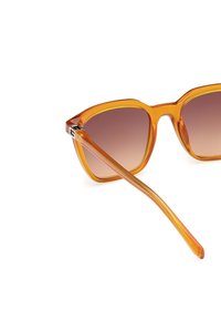 Gafas de sol transparentes naranjas con un marco geométrico, lentes marrones degradados y brazos delgados. Acentos de hardware notables en el marco.