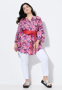 Bloemen tuniek in roze en paars met lange mouwen, cinched in de taille met een rode riem, gedragen over witte slim fit broek en zwarte sandalen.