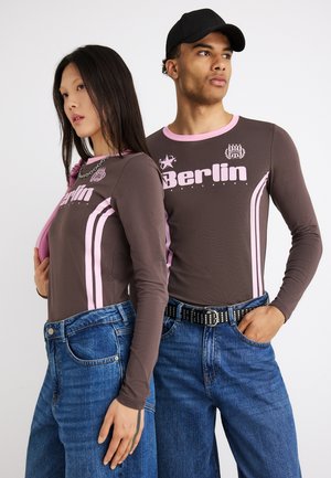 Dwa modele noszące dopasowane brązowe topy z długim rękawem z różowym napisem „Berlin” i paskami, zestawione z niebieskimi dżinsami; jeden model nosi czapkę z daszkiem w kolorze czarnym.