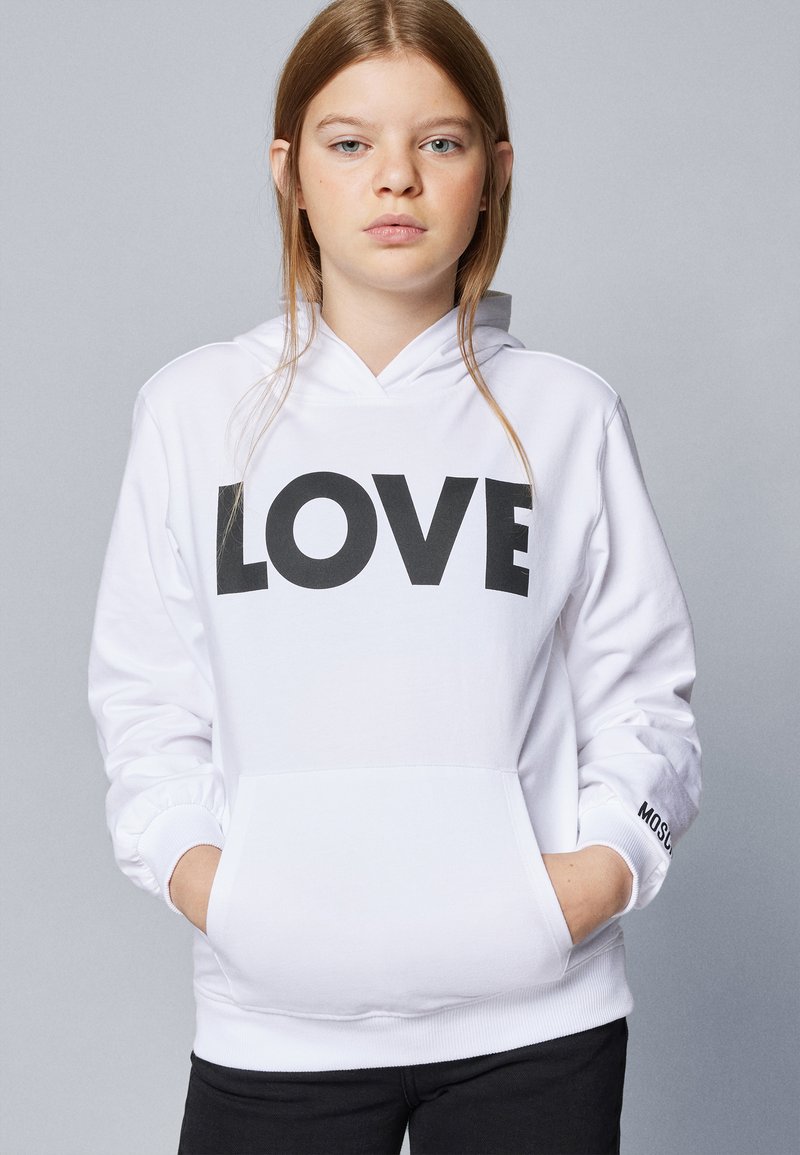Hvit hettegenser laget av bomull, med et stort svart "LOVE" trykk på forsiden, en frontlomme og ribbestrikkede mansjetter med svart tekstakcent.