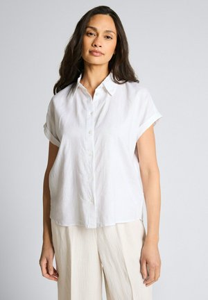 Kobieta z długimi ciemnymi włosami, ubrana w luźną białą koszulę na krótki rękaw z guzikami oraz jasnobeżowe spodnie, stoi na jednolitym tle.