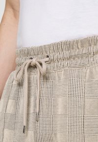 Pantalons beiges à motif avec une taille élastique, un détail de cordon de serrage et divers motifs de grille texturés en noir et beige.