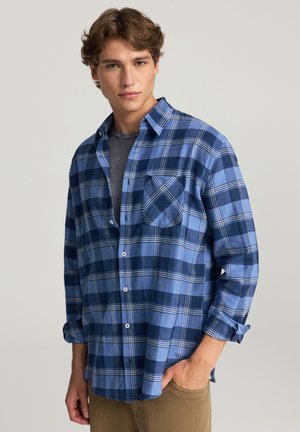 Camicia in flanella a quadri blu con chiusura a bottoni, taschino sul petto, e maniche lunghe, indossata sopra una maglietta grigia. Tessuto morbido.