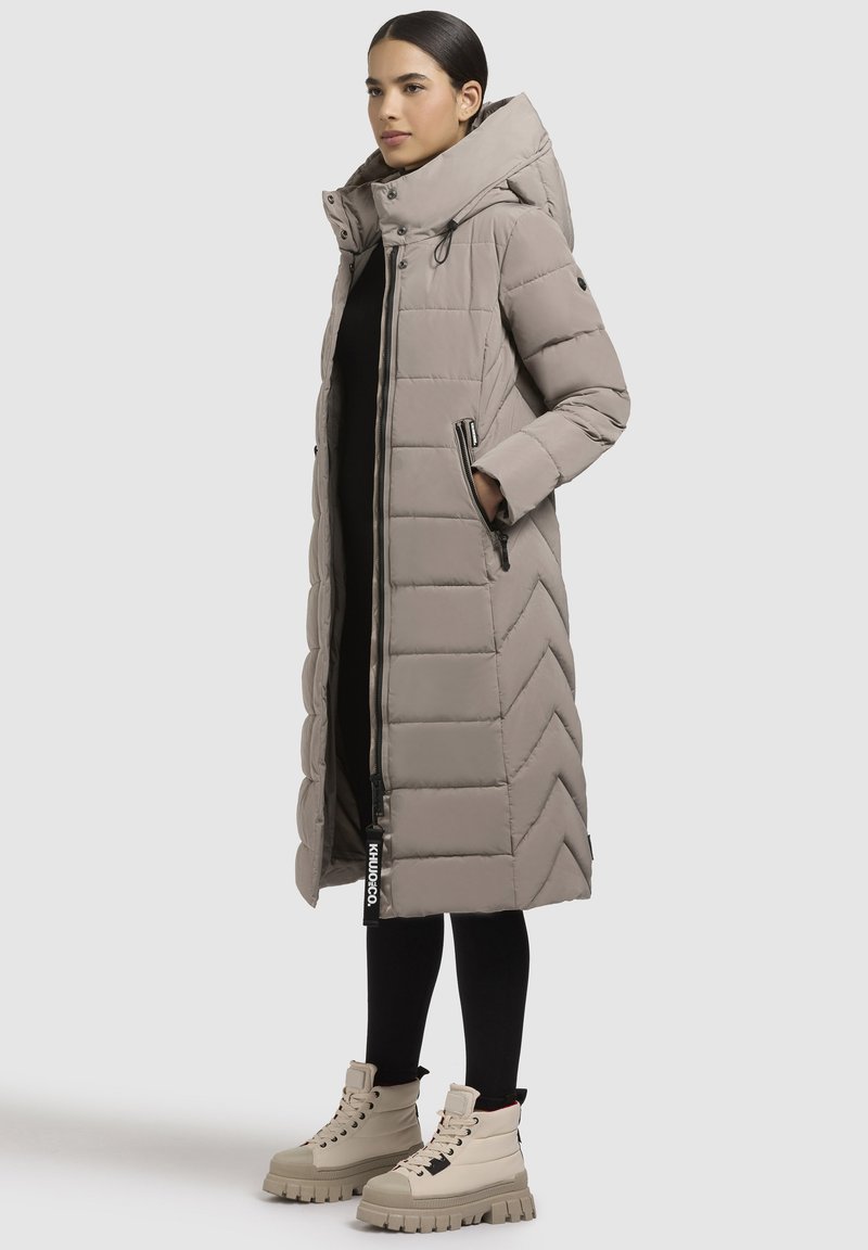 Woman wearing a long taupe quilted puffer coat with hood, black leggings, and beige chunky lace-up boots, standing against a plain background.