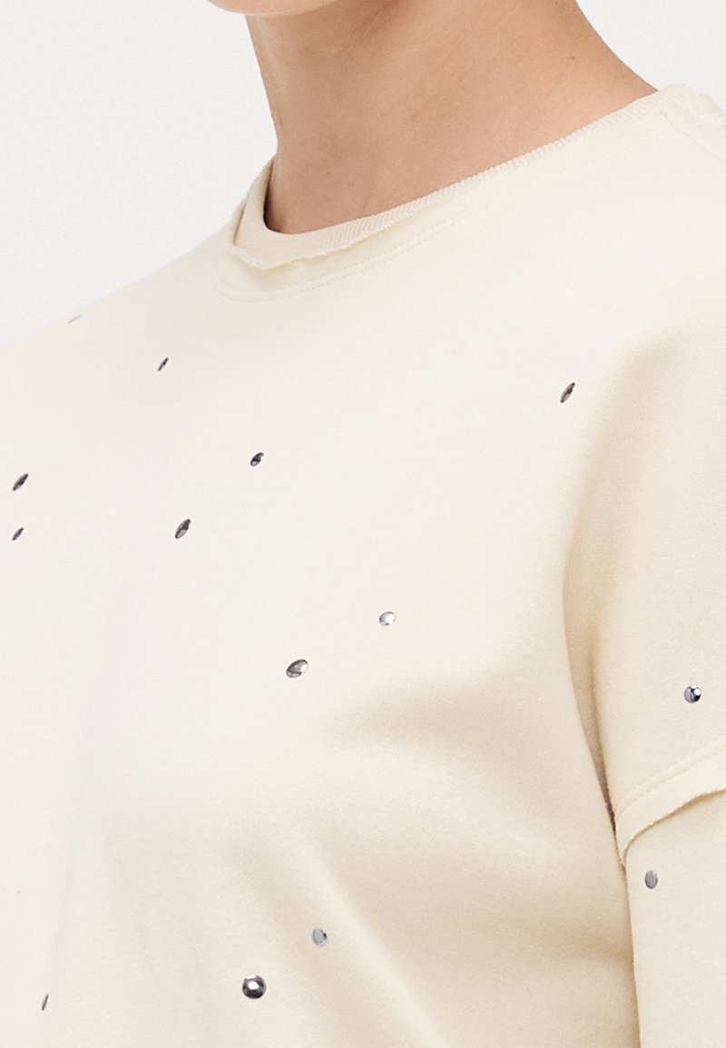 Gros plan d'une personne portant un sweat-shirt crème décoré de petits clous brillants dispersés.