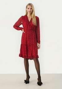 Rode bloemenjurk met lange doorzichtige mouwen, v-hals en rimpelzoom. Gepaard met zwarte leggings en schoenen met grote strikken.