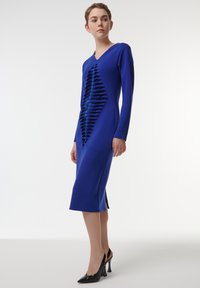 Vestido de manga larga de un azul vibrante, que presenta un patrón texturizado en zigzag en la parte delantera y un escote en V. Combinado con zapatos de tacón alto negros.