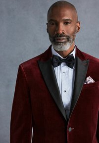 Burgundska baršunasta tuxedo jakna s crnom satenskom reverom, uparena s bijelom košuljom i crnim leptir mašnom. Ima uzorčanu maramicu.