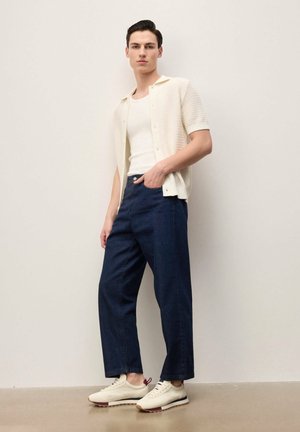 Young man standing against a white wall wearing a white short-sleeve knit shirt, white tank top, dark blue wide-leg pants, and white sneakers.