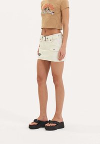 Mini gonna in denim bianco con vita con cintura, tasche laterali e dettagli ricamati neri. Abbinata a una maglietta cropped color sabbia e sandali platform neri.