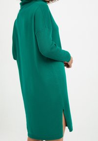 Robe-pull verte avec un col montant, des manches côtelées et des fentes sur les côtés. Le tissu doux présente une texture unie avec une coupe décontractée.