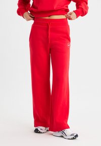 Rote Sweatpants mit einem Kordelzugbund, weiter Beinform und seitlichen Taschen. Glatte Stoffstruktur mit einem kleinen Logo auf dem Oberschenkel.