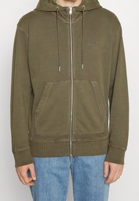 Sudadera con cremallera en verde oliva hecha de tela suave, con capucha, dos bolsillos delanteros y puños y bajo acanalados. Cremallera y cordones plateados.