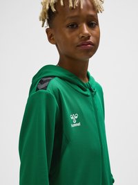 Chłopiec z blond dredami w zielonej bluzie z kapturem zapinanej na zamek, z białym logo hummel na piersi, na jednolitym tle.