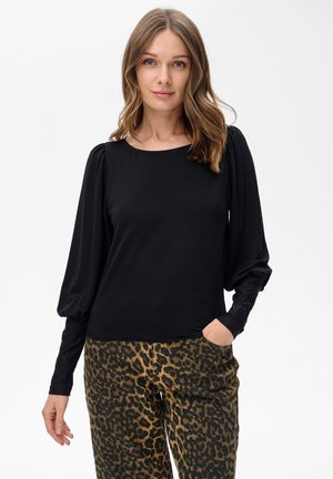 Kobieta z luźnymi falującymi włosami w czarnej bluzce z długimi rękawami i bufkami oraz spodniami w panterkę, stojąca na białym tle.