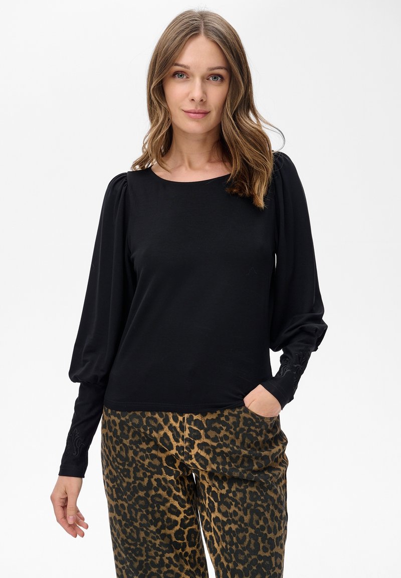 Kobieta z luźnymi falującymi włosami w czarnej bluzce z długimi rękawami i bufkami oraz spodniami w panterkę, stojąca na białym tle.