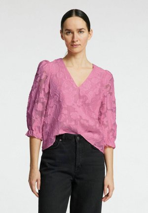 Lyserød bluse med blomsterstruktur, V-udskæring og puffede ærmer, parret med sorte højtaljede jeans. Blødt, gennemsigtigt stof med lagdelte detaljer.