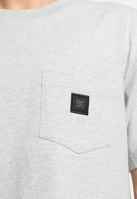 Grå bomulds T-shirt med rund hals, der har en forlommeslomme og et lille sort logo mærke. Blød tekstur, kortærmet.