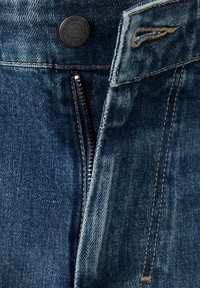 Gros plan de la taille d’un jean en denim bleu avec un bouton noir étiqueté "MNG DENIM" et une braguette partiellement fermée.