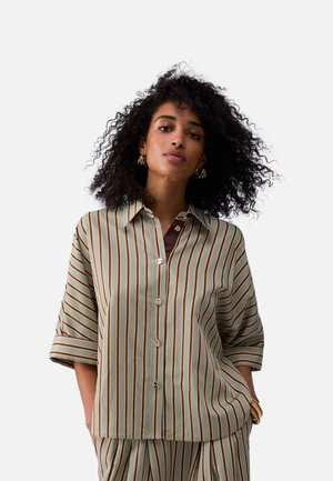 Mujer con cabello rizado vistiendo una camisa de botones beige y marrón con rayas verticales y pantalones a juego, posando con las manos en los bolsillos.