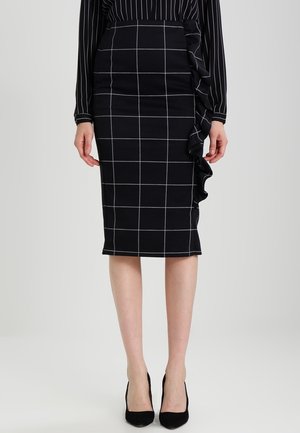 Femme portant une jupe crayon noire à lignes blanches en grille et volant latéral, associée à un chemisier noir à manches longues avec des rayures verticales blanches.