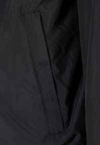 Svart ytterkläder med en slät yta, en sidficka och rena söm detaljer. Tyget verkar lätt och hållbart.