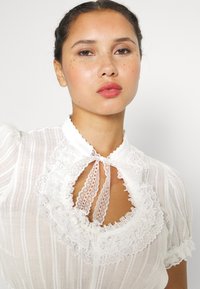 Λευκό πουκάμισο με κολλέ στην δαντέλα και λεπτομέρεια με κορδόνι, με κάθετες ρίγες και υφασμάτινη υφή, φουσκωτά μανίκια και κλειδί κοπής.