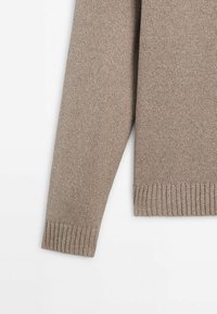 Manche de pull beige tricoté avec détails de bord-côte aux poignets et à l'ourlet sur fond blanc.