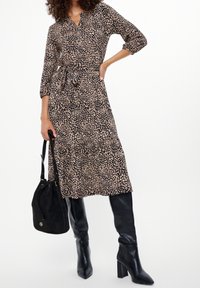 Černobéžové šaty s vzorem, výstřihem do V, tříčtvrtečními rukávy a zavazováním v pase, doplněné černými vysokými botami ke kolenům a černou kabelkou.
