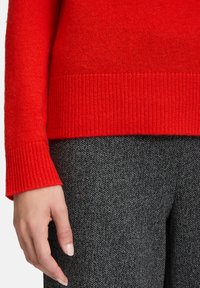 Osoba ubrana w czerwoną wełnianą sweter i szare spodnie w wzór herringbone, z lewą ręką luźno opisaną przy boku.