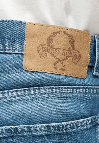 Ανοιχτό μπλε τζιν παντελόνι με ταμπόν από ταμπά δέρμα στη ζώνη, με ανάγλυφη επιγραφή «Moschino Milano» και σχέδιο δάφνινου στεφάνου.