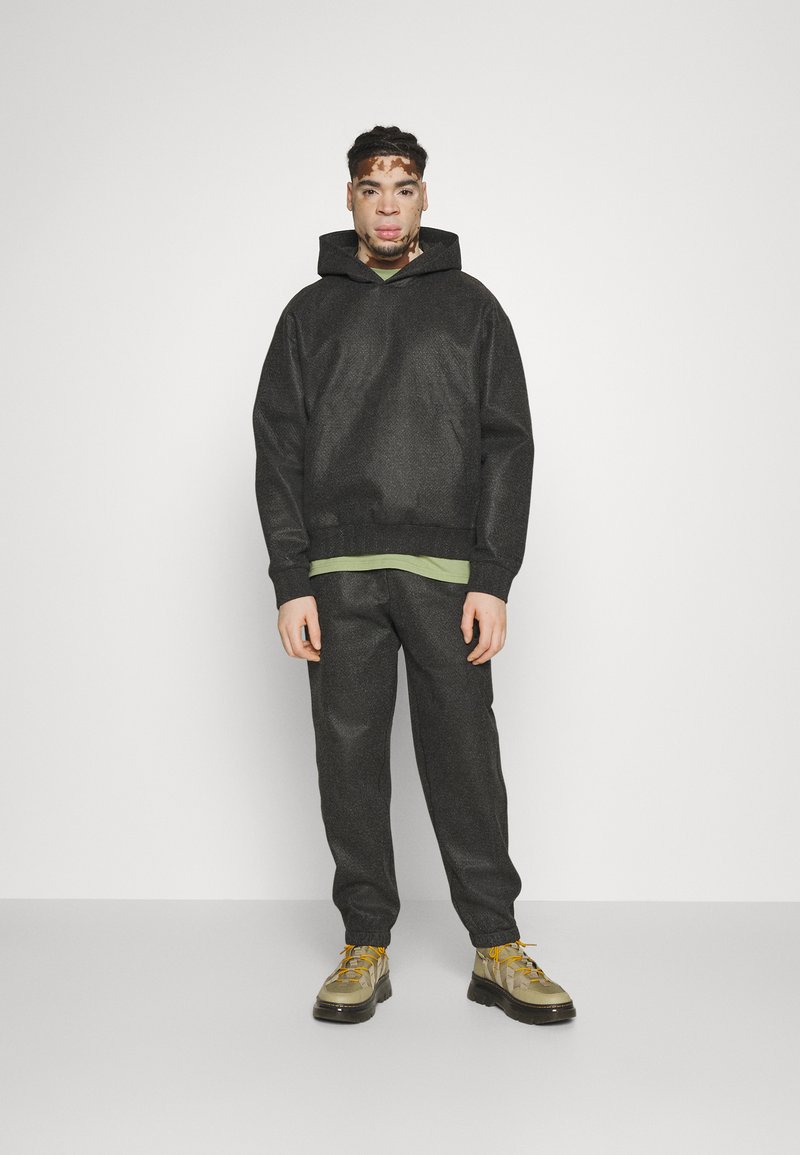 Un sweat à capuche noir texturé avec un pantalon assorti, superposé sur une chemise verte. L'ensemble est complété par des bottes de randonnée beiges, avec des lacets jaunes.