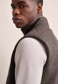 Braunes Fischgrät-Weste mit hohem Kragen über einem gerippten, hellen Pullover. Glatte Textur, taillierte Passform und kontrastierender dunkler Besatz.