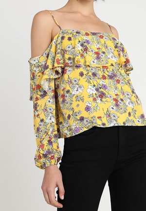 Top giallo off-the-shoulder con motivo floreale, volant stratificati e maniche lunghe con polsini elasticizzati. Il materiale sembra leggero e fluido.
