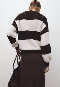 Suéter de punto a rayas marrón y blanco con un corte holgado y puños acanalados, combinado con una falda marrón oscuro. Los accesorios incluyen un bolso negro.