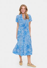 Blauw en wit gedessineerde jurk met korte mouwen, knoopsluiting en een vloeiende rok. Draagt met beige platgenaaide schoenen. Glad stofoppervlak.