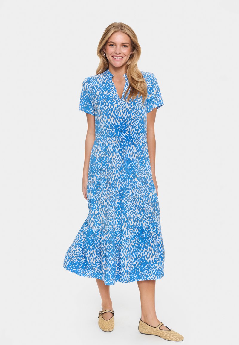 Blauw en wit gedessineerde jurk met korte mouwen, knoopsluiting en een vloeiende rok. Draagt met beige platgenaaide schoenen. Glad stofoppervlak.