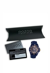 Montre en silicone bleu avec un cadran rond, des accents dorés et des index d'heure proéminents. Emballage noir avec le logo "oozoo timepieces" et une carte de garantie.