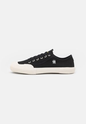 Black canvas sneakers with white rubber sole, round toe, lace-up design, circular eyelets, and small white logo on the side.
