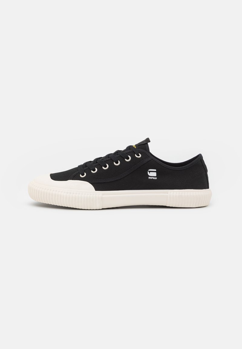 Zapatillas de lona negras con suela de goma blanca, punta redonda, diseño con cordones, ojales circulares y pequeño logo blanco en el lado.