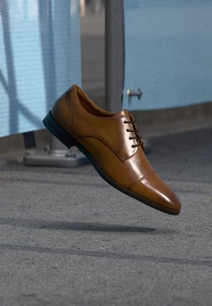 Une chaussure en cuir marron cirée à lacets flotte au-dessus d'un trottoir gris, avec un mur à panneaux bleus derrière.